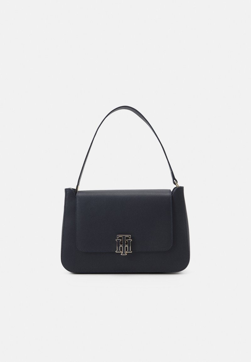 Sac à main en cuir noir avec une poignée supérieure unique et un fermoir logo en métal doré sur le rabat avant sur un fond clair.