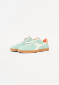 Light green suede sneakers with a white logo, orange accents on the heel and tongue, and a gum rubber outsole. Classic lace-up design.