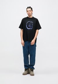 Svart t-skjorte med korte ermer og grafisk design på forsiden, kombinert med løse blå jeans og olivengrønne støvler.