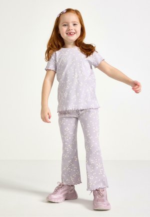 Smilende rødhåret pige iført en matchende lys lilla blomsterprintet skjorte og bukser med lyserøde sneakers, stående med den ene arm udstrakt.