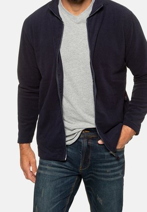 Homme portant une veste zippée bleu marine sur un t-shirt gris et un jean bleu, avec une main dans la poche et l'autre détendue le long du corps.