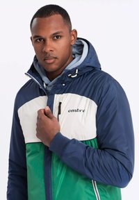 Färgglad jacka med marinblå, vit och grön paneler. Inkluderar dragkedjestängning, justerbar huva och "ombre"-logotyp på bröstet.