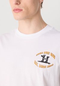 T-shirt in cotone bianco con una grafica circolare in nero e arancione. La stampa include i dettagli "HUGO SURF CLUB" e "EST. SINCE 1993".