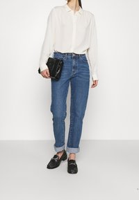 Camicia bianca a maniche lunghe, jeans blu a gamba dritta, borsa nera e mocassini neri con dettagli in catena argentata, su uno sfondo neutro.