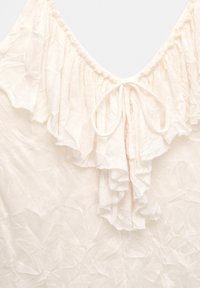 Off-white tyg med volangkrage och rosett, med en subtil blommönsterstruktur på det lätta materialet.