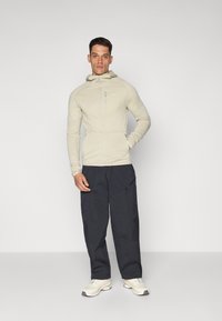 Haut à capuche zippé beige clair avec un motif texturé, doté d'une capuche et d'une poche poitrine, associé à un pantalon cargo ample et sombre.