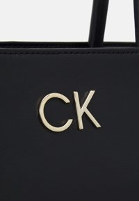 Czarna torebka wykonana z teksturowanego materiału, z akcentem złotego logo „CK”. Prosty design z krótkimi uchwytami, podkreślający nowoczesną elegancję.