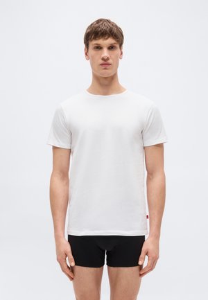 Mladý muž stojaci čelom vpredu, oblečený v jednoduchom bielom tričku s krátkym rukávom a čiernych boxerkách, na bielom pozadí.