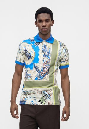 Homme portant un polo à manches courtes avec un col bleu et un imprimé multicolore représentant des paysages urbains et des motifs abstraits.