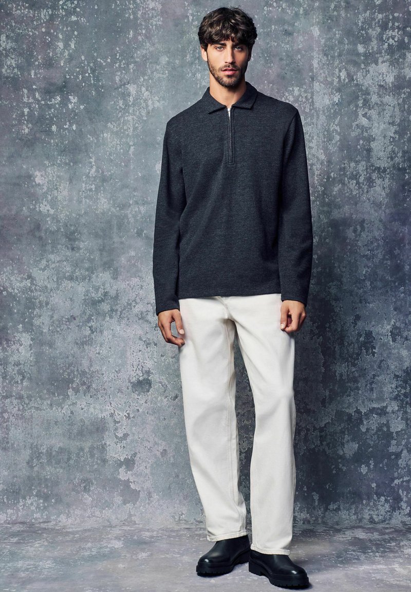 Donkergrijze lange mouwen polo met een ritskraag, gecombineerd met witte rechte broek en zwarte robuuste laarzen. Zachte stof, minimalistisch ontwerp.