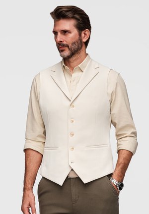 Homme barbu portant un gilet crème sur une chemise à manches retroussées et un pantalon olive, debout avec une main dans la poche, fond neutre.