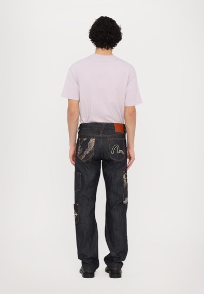 Chemise courte à manches courtes lavande avec un jean bleu foncé arborant divers patchs et des coutures orange, avec un accent en cuir texturé sur la taille.