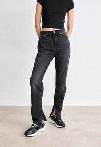 High-waisted black denim jeans with a straight leg, featuring a split hem detail at the ankle. Paired with black and white sneakers.