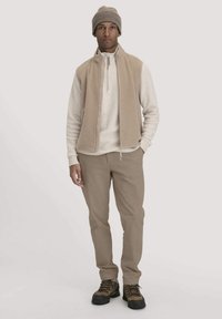 Mann trägt eine beige Strickmütze, eine beige Fleeceweste mit Reißverschluss, ein helles Langarmshirt, khakifarbene Hose und braune Wanderschuhe und steht vor einem weißen Hintergrund.