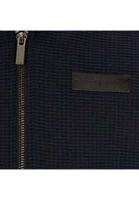 Svart och marinblått rutigt mönster på tyg med en läderlabel och silverfärgad dragkedja. Texturen är vävd, med vertikala och horisontella ränder.