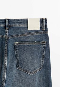 Jean en denim bleu avec une étiquette en cuir blanc propre sur la taille et une poche arrière unique ornée d'un motif de couture subtil.