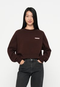 Felpa corta marrone con logo "Tommy" rosa. Presenta un collo rotondo e maniche leggermente bombate, abbinata a jeans scuri.
