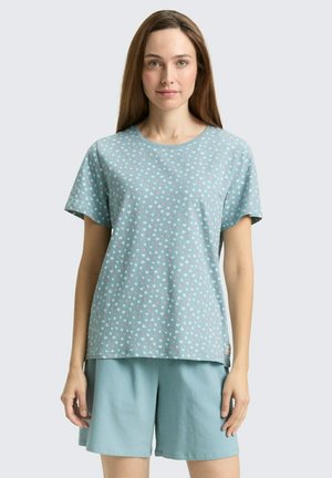 Vrouw draagt een lichtblauw kortermig shirt met kleine hartjespatronen en bijpassende effen lichtblauwe shorts, staande tegen een effen achtergrond.