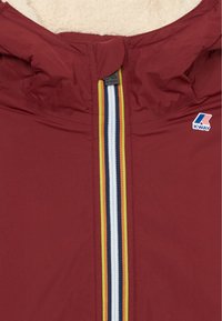 Bordeauxjakke med fleece-fôret krage, sentral glidelås med hvite, marineblå, oransje og gule striper, K-Way-logo på brystet.