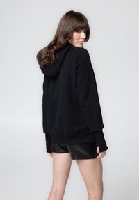 Czarna bluza z kapturem, wykonana z miękkiego, teksturowanego materiału, z okrągłym kapturem i ściągaczami przy mankietach. Noszona z czarnymi szortami, prezentując swobodny styl.