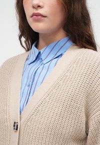 Kvinne iført en blåstripet skjorte med krage under en beige tykk strikkegenser med en stor knapp, vist fra haken til midten av brystet.