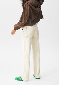 Luźne kremowe spodnie z widocznymi przeszyciami, zestawione z brązową krótką kurtką z kapturem i zielonymi sandałami z odkrytymi palcami.