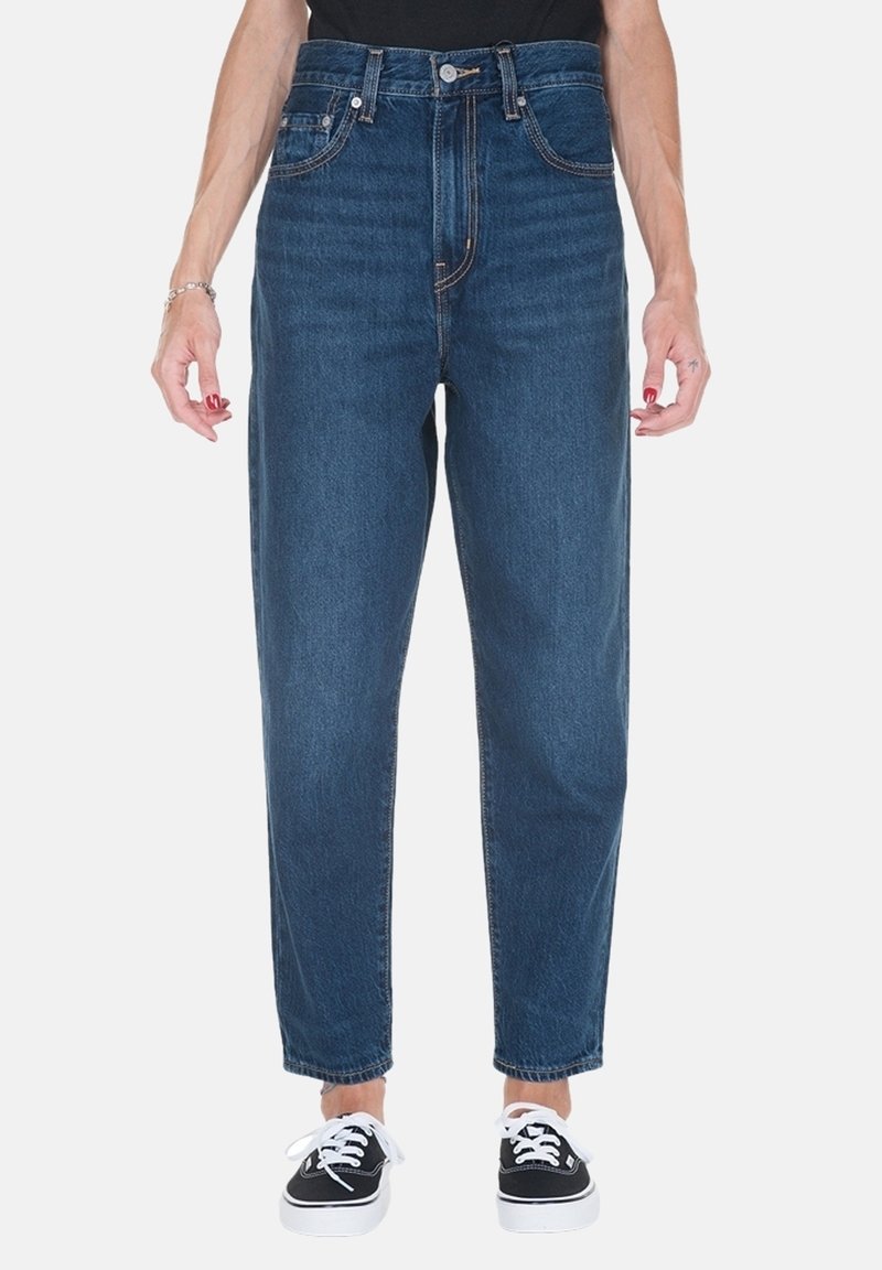 Jean taille haute bleu foncé avec une coupe droite, design à cinq poches et coutures contrastantes, associés à des baskets noires.