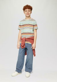Gestreept T-shirt in groen, blauw, bruin en oranje, gecombineerd met lichtblauwe wijde jeans en een roestkleurige trui om de taille gebonden.