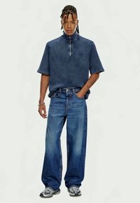 Navy-blaues Cord-Hemd mit kurzen Ärmeln und Reißverschlusskragen, kombiniert mit hellen, weit geschnittenen Denim-Jeans. Graue Sneakers runden den Look ab.