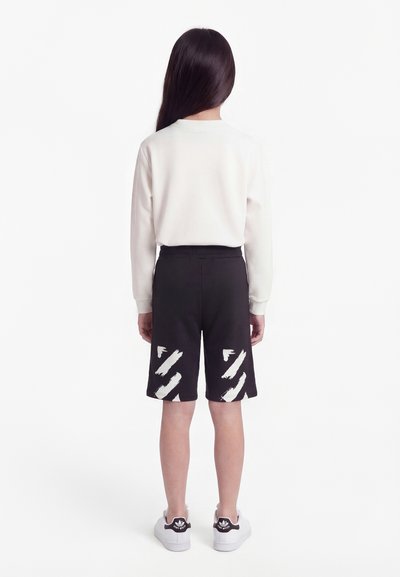 Παιδί με μακριά σκούρα μαλλιά, που στέκεται με την πλάτη, φορώντας λευκό πουκάμισο με μακριά μανίκια, μαύρες σορτς με λευκά σχέδια με πινελιές και λευκά αθλητικά παπούτσια.