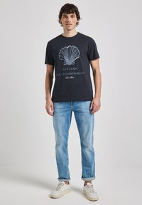 Černé bavlněné tričko s bílou grafikou mušle a textem "BLUE CLAM BAR AND RESTAURANT" v neformálním písmu. V kombinaci se světlými modrými džíny.