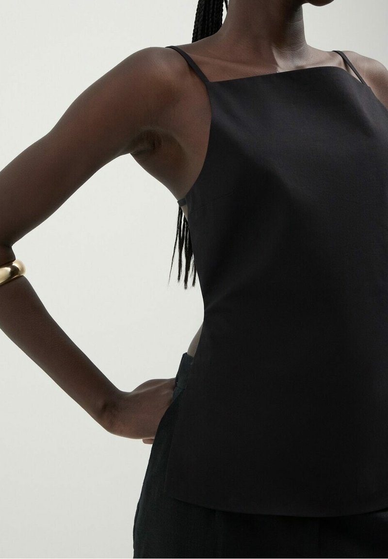 Femme portant un haut noir sans manches à fines bretelles, debout avec une main sur la hanche et un bracelet en or au bras.
