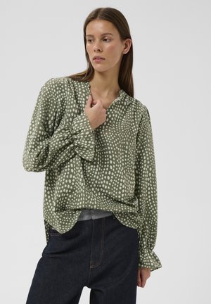 Vrouw draagt een olijfgroene blouse met witte stippen, gerimpelde manchetten en donkere blauwe jeans, kijkt naar links tegen een effen achtergrond.