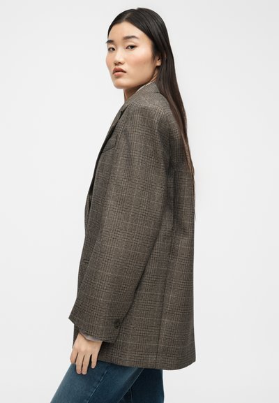 Γυναίκα με μακριά σκούρα μαλλιά, φοράει καφέ καρό σακάκι και μπλε τζιν, κοιτώντας ελαφρώς πάνω από τον ώμο της, με φόντο απλό.