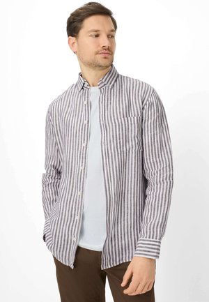 Man draagt een wit T-shirt onder een lang mouw overhemd met verticale grijs-witte strepen, bruine broek, handen in de zakken.