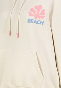 Beige Kapuzenpullover mit einer rosa Muschel-Grafik und blauer "BEACH"-Schrift. Verfügt über eine Fronttasche und Kordeln mit rosa Tipps. Weicher Stoff.