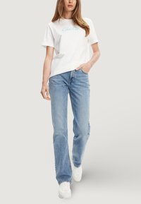 Donna in piedi che indossa una t-shirt bianca Calvin Klein, jeans azzurri e sneaker bianche, con una mano in tasca, su sfondo semplice.