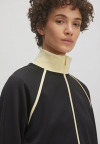 Zwarte ritssluitingjas met een hoge kraag, voorzien van crèmekleurige accenten langs de schouders en de rits, gemaakt van een soepel stof.