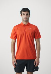 Nike polo skjorte i lys orange, korte ermer, klassisk krage, med tre-knappers stolpe, med en hvit swoosh-logo på brystet.
