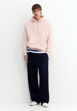 Un jeune homme se tient contre un mur blanc, portant un sweat à capuche rose pâle, un pantalon large bleu marine, une chemise rayée en dessous et des chaussures de couleur claire.