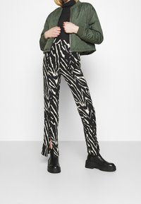 Pikowana zielona kurtka bomberka zestawiona z czarnymi spodniami w kremowo-czarży wzór zebry z rozcięciami oraz masywnymi czarnymi platformami za kostkę.