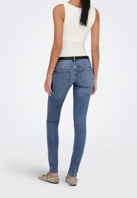 Femme debout, dos visible, portant un haut blanc sans manches, un jean skinny bleu et des chaussures plates à motif serpent.