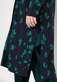 Stickad cardigan i marinblått med grönt blommönster, med sidofransar och långa ärmar. Tyget verkar mjukt med en texturerad finish.