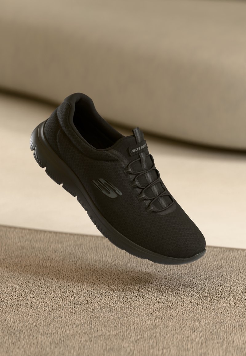Black Skechers shoe with a mesh upper, textured finish, featuring elastic laces and a cushioned sole, suspended above a textured surface.