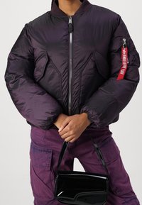 Fioletowa kurtka puchowa z zamkiem błyskawicznym, prążkowanym kołnierzem i oversize'owymi rękawami. Zawiera metkę "Remove Before Flight". W zestawie z czarną torbą.