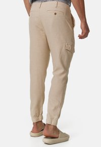 Beige linen pants with elastic cuffs, featuring two back pockets and a side cargo pocket. Smooth texture with a casual fit.