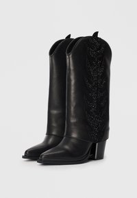 Black leather cowboy boots with pointed toes, high shafts, and decorative leaf-shaped accents featuring rhinestones. Block heels.