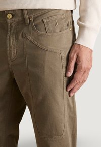 Pantalones caqui con corte recto, fabricados en un algodón resistente, con bolsillos frontales con detalles de costura y un logo de marca visible en el bolsillo.