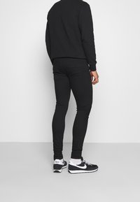 Černá mikina a slim-fit černé džíny s hladkým povrchem. Outfit dotvářejí černobílé tenisky, pohled ze zadu.