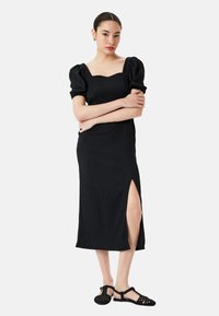 Vestido negro de canalé, hasta la rodilla, con mangas cortas abullonadas y escote cuadrado. Presenta una abertura lateral como detalle adicional. Se lleva con sandalias negras.